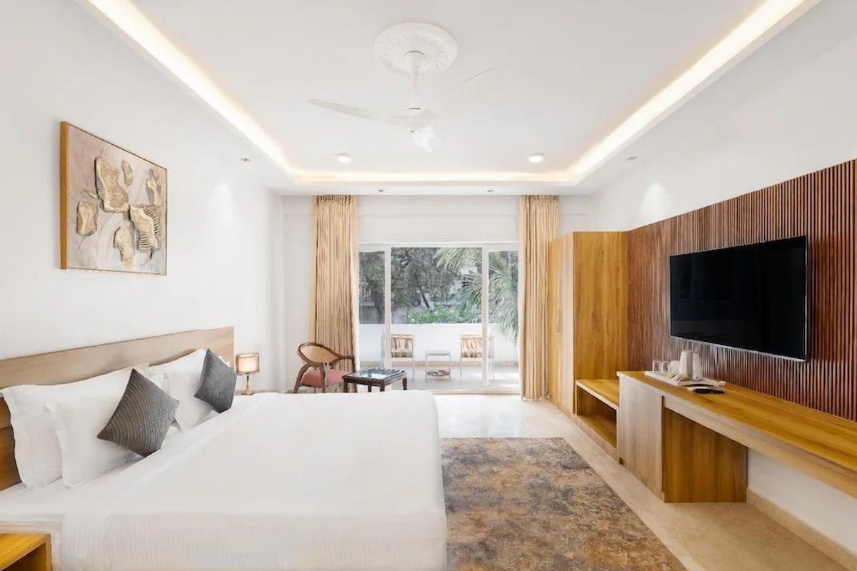 A modern hotel room with a large bed, a flat-screen TV mounted on a wooden panel, a ceiling fan, and a view of the outdoors through large windows.
