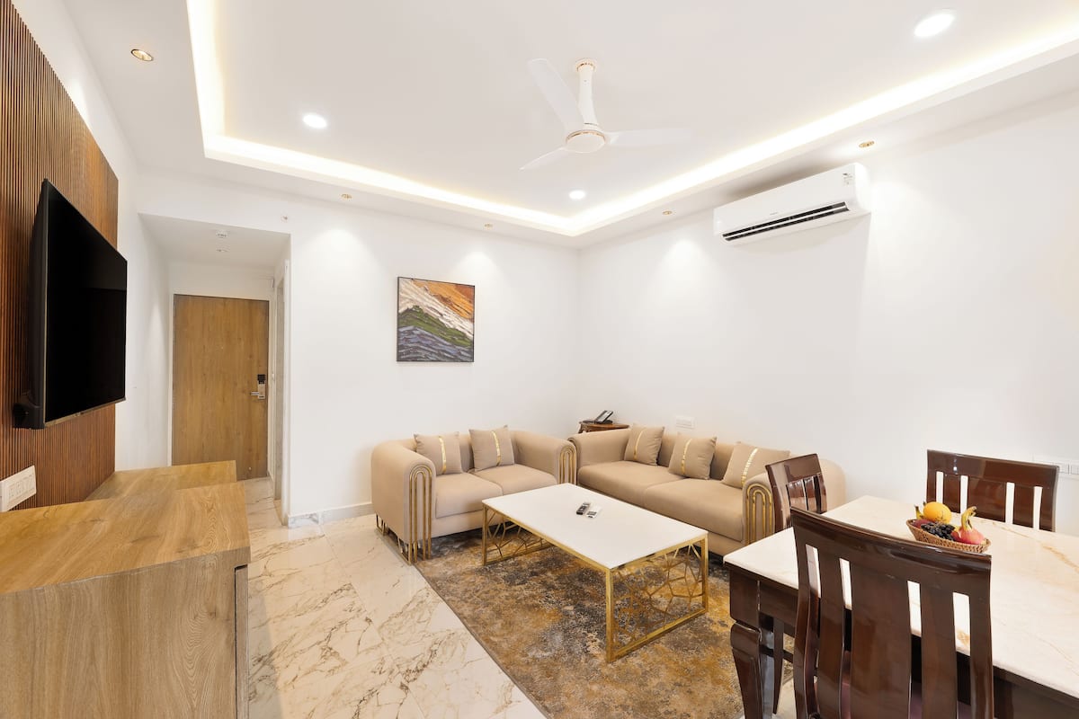 A modern living room with a beige sectional sofa, a white coffee table, and a dining table with chairs. There is a flat-screen TV mounted on the wall, a ceiling fan, and recessed lighting.