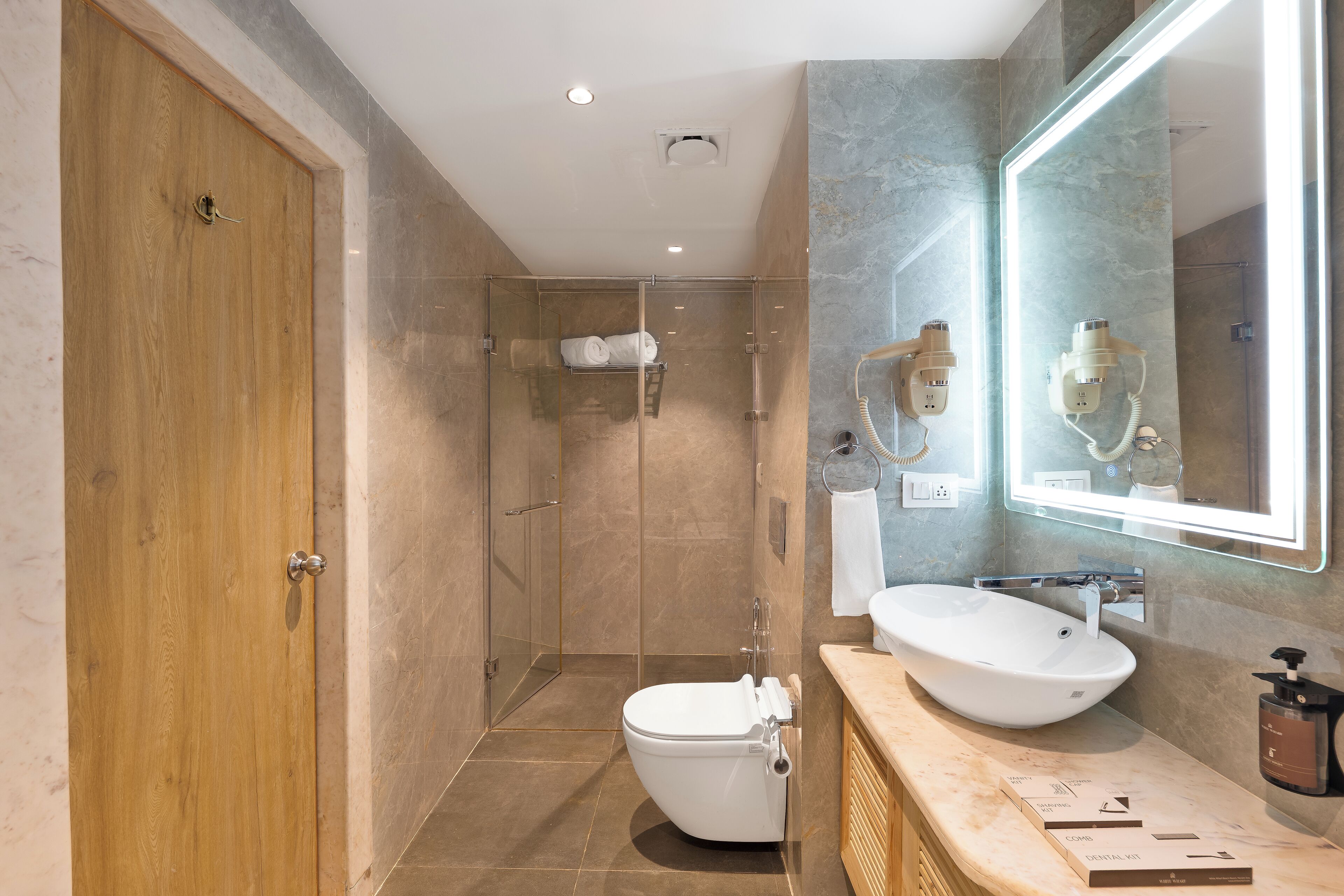 A modern bathroom with a wooden door, a glass-enclosed shower, a white sink, and a large mirror.