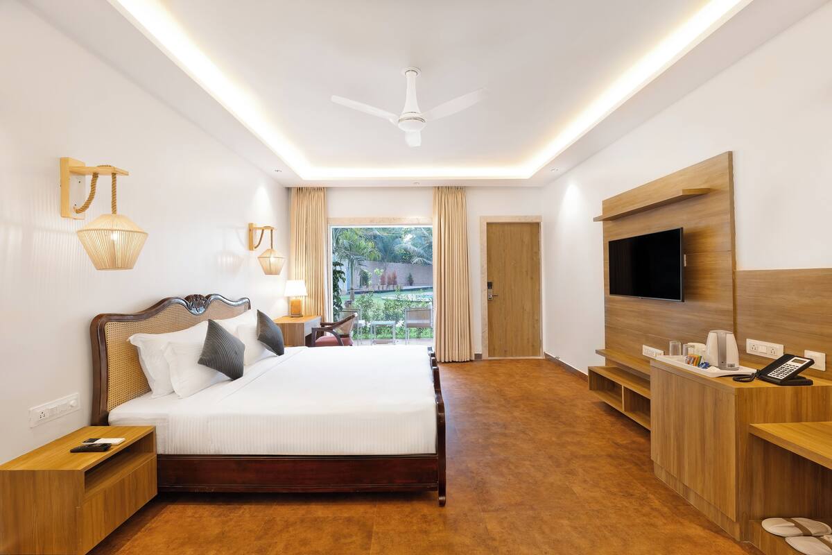 A hotel room with a bed, bedside tables, a TV, a desk with a phone, and a view of greenery outside.