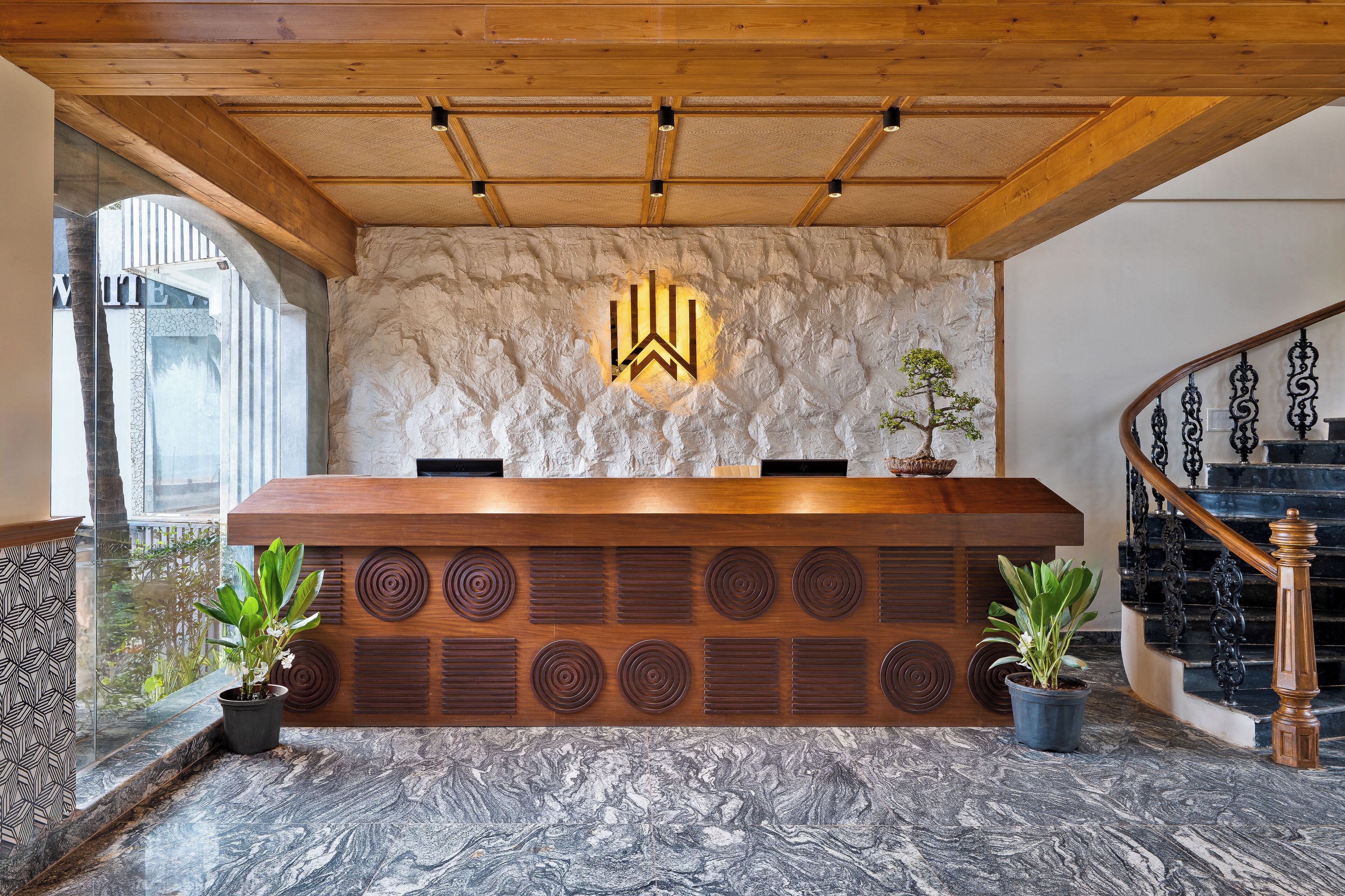 A reception desk with a wooden finish, a stone wall with a logo, and a staircase with black railings.