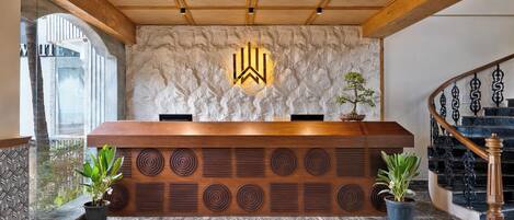 A reception desk with a wooden finish, a stone wall with a logo, and a staircase with black railings.