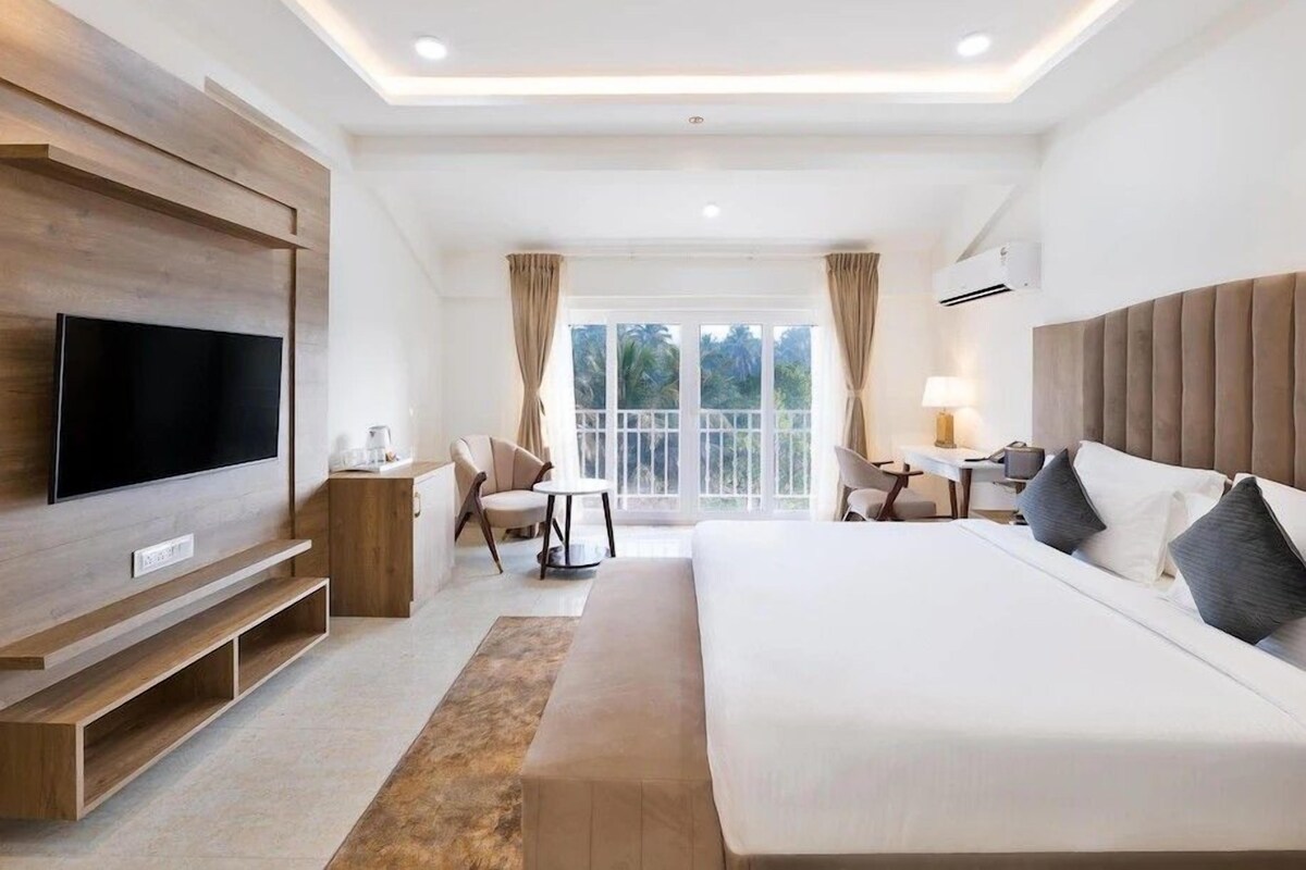 A modern hotel room with a large bed, a flat-screen TV mounted on the wall, a small desk with a lamp, and a view of greenery through the windows.