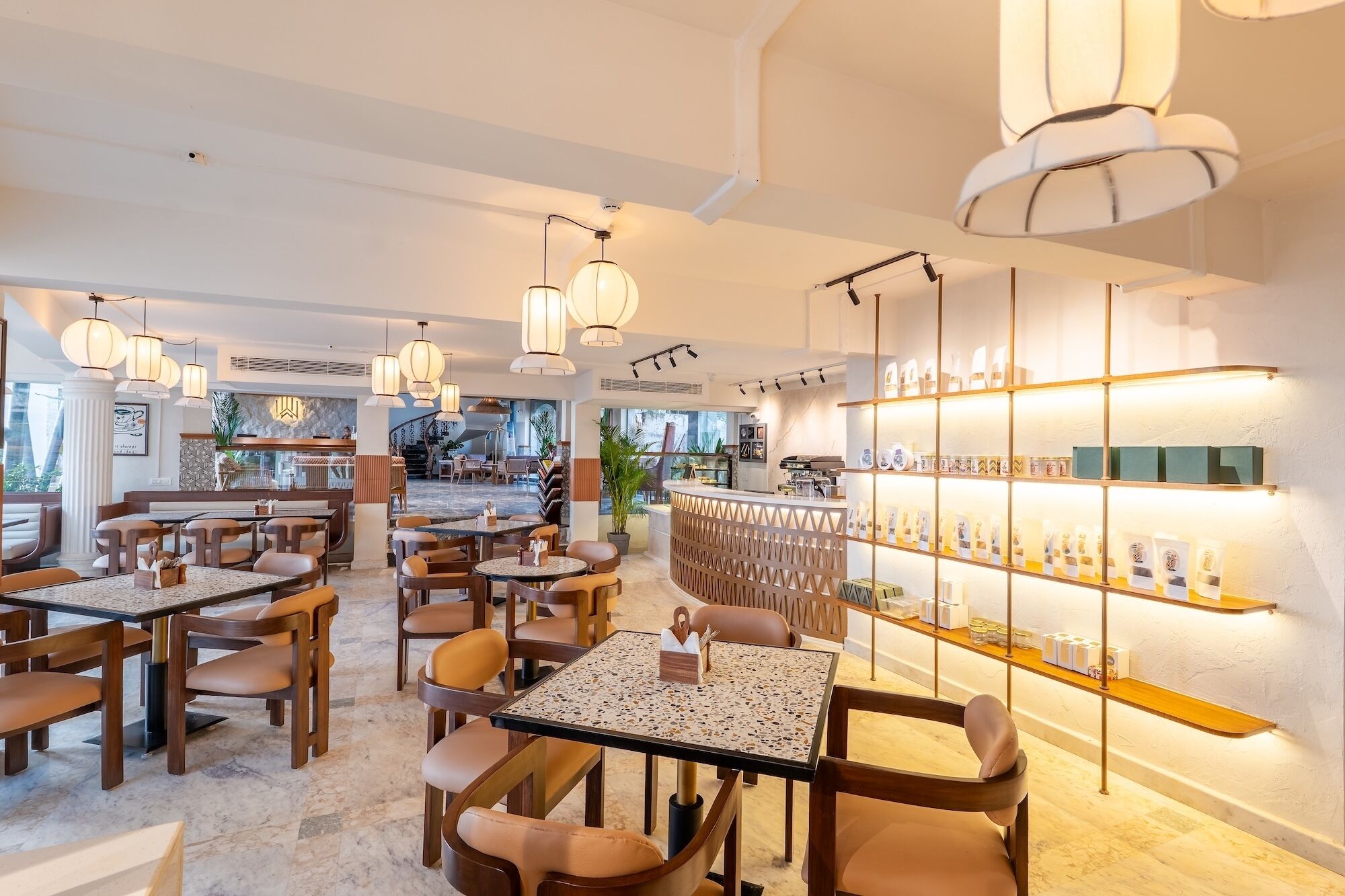 A modern restaurant interior with multiple dining tables and chairs, pendant lighting, and a bar area with shelves displaying items.