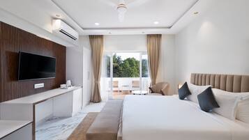 A modern hotel room with a large bed, a flat-screen TV, a desk, and a balcony with a view of greenery.