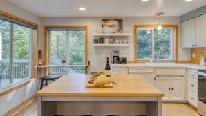 A modern kitchen with a central island, wooden cabinetry, and large windows offering views of greenery. - Arch Cape Retreat • Hot Tub • Near Cannon Beach + Haystack Rock | Royal Canoe (Arch Cape)