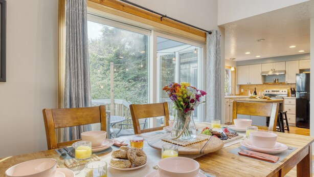 A breakfast table set with pink dishes, a vase of flowers, and a view of the kitchen. - Arch Cape Retreat • Hot Tub • Near Cannon Beach + Haystack Rock | Royal Canoe (Arch Cape)