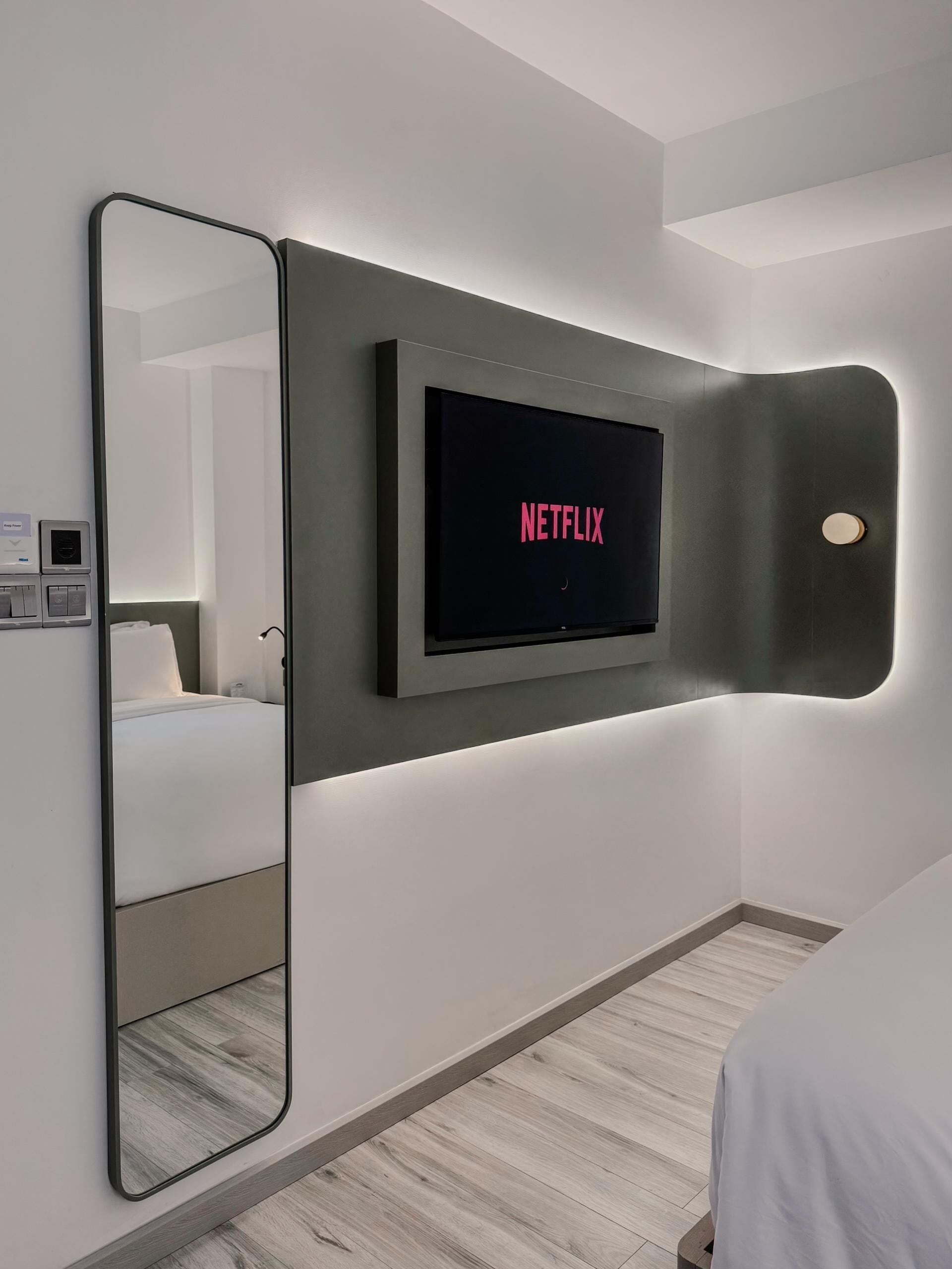 Een moderne hotelkamer met een grote flatscreen televisie aan de muur, een spiegel in volle lengte en een bed met een witte deken.
