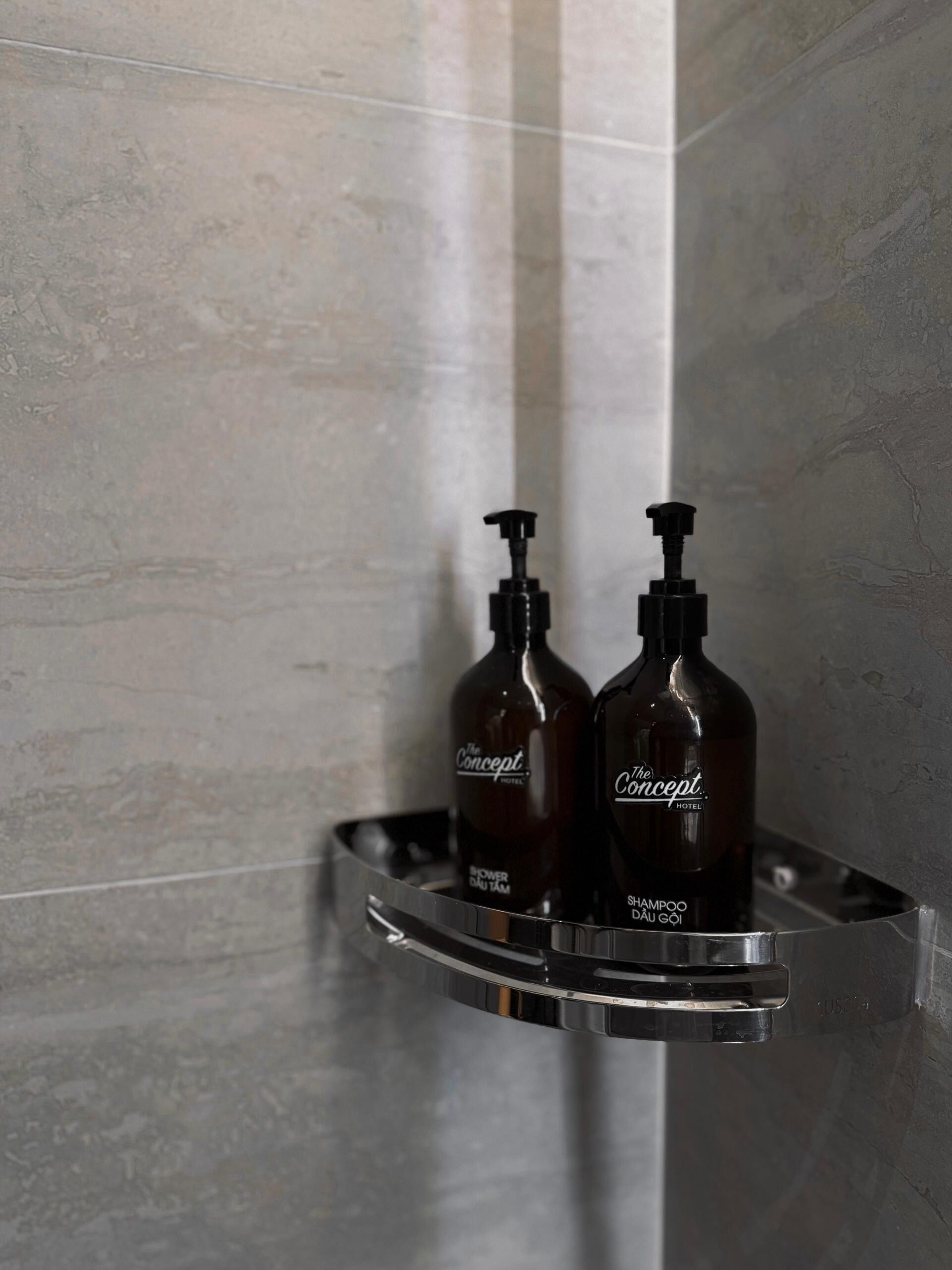 A bathroom with a shower area, a metal shelf holding two bottles of The Concept Hotel shower gel, and a textured wall.