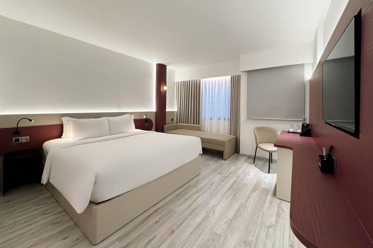 A modern hotel room with a large bed, a desk, a chair, a TV, and a window with curtains.
