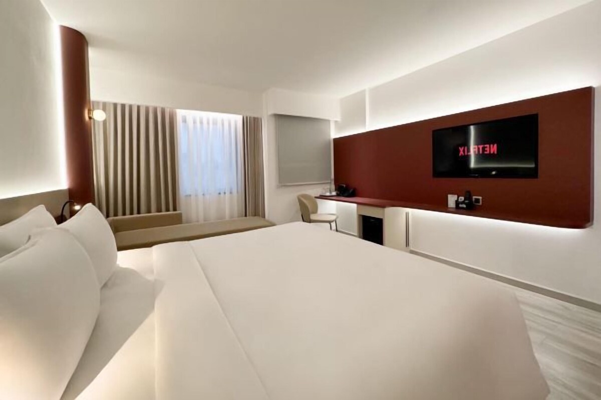 A modern hotel room with a large bed, a desk with a flat-screen TV, and a seating area with curtains.