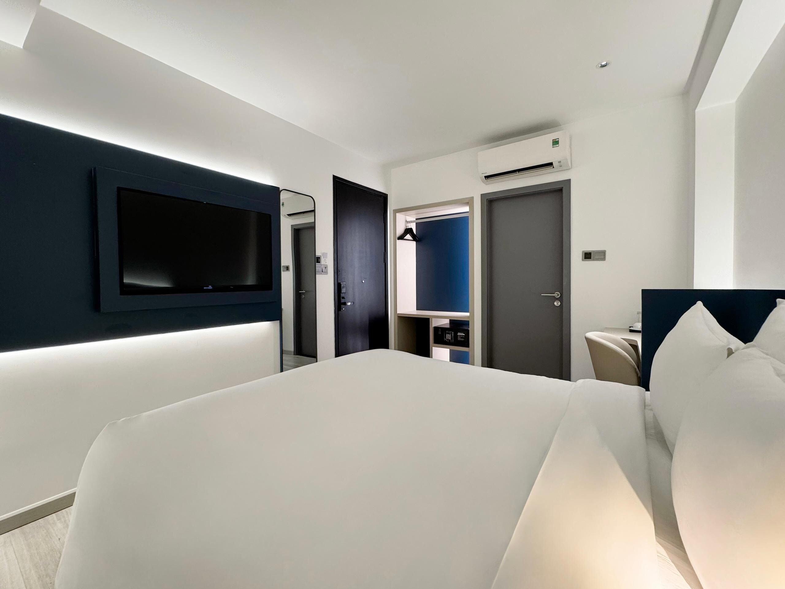 A modern hotel room with a large bed, a flat-screen TV mounted on the wall, a wardrobe, and an air conditioning unit.