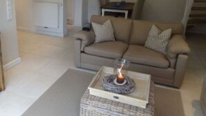 A living room with a beige sofa, a small table with a candle, and a kitchen area in the background. - Holiday Home 'Sylthus-Haithabu' with Private Terrace, Private Garden and Wi-Fi (Wenningstedt)