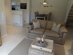 A living room with a beige sofa, a small table with a candle, and a kitchen area in the background.