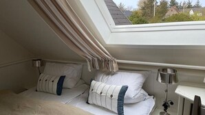 A bedroom with a skylight, a bed with white bedding, two pillows, a bedside table with a lamp, and a view of trees and houses outside. - Holiday Home 'Meerchenhaus' with Private Terrace, Garden, and Wi-Fi (Wenningstedt)