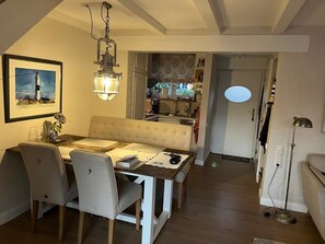 Un espace repas comprenant une table, des chaises, un banc, une suspension, un tableau encadré et, au fond, une cuisine.