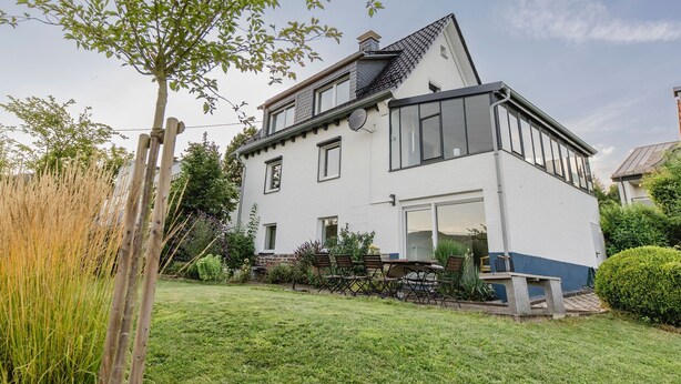 Una casa de dos pisos con fachada blanca, un balcón y un jardín con cerca de madera y un árbol.