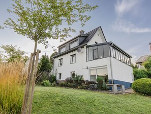 Una casa de dos pisos con fachada blanca, un balcón y un jardín con cerca de madera y un árbol.