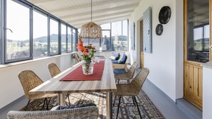 Un área de comedor con una mesa, sillas, un florero con flores, una lámpara colgante y grandes ventanales con vistas.