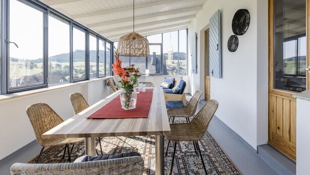 Un área de comedor con una mesa, sillas, un florero con flores, una lámpara colgante y grandes ventanales con vistas.
