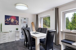 A modern dining area with a white table, black chairs, and a bench. There is a painting on the wall, a window with a view of trees, and a radiator. - Apartment 'Villa Eichwald' with Shared Garden, Balcony and Wi-Fi (Freudenstadt)