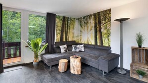 A modern living room with a grey sectional sofa, a large forest mural, a tall floor lamp, and a wooden coffee table. - Apartment 'Villa Eichwald' with Shared Garden, Balcony and Wi-Fi (Freudenstadt)