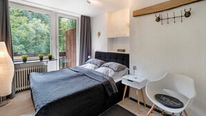 A modern bedroom with a bed, bedside table, chair, and a view of greenery outside. - Apartment 'Villa Eichwald' with Shared Garden, Balcony and Wi-Fi (Freudenstadt)
