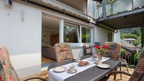 An outdoor dining area with a table set for two, surrounded by patterned chairs, a view of the interior through a glass door, and a balcony with a railing. - Apartment 'Design Und Entspannug' with Private Terrace, Private Garden and Wi-Fi (Kandern)