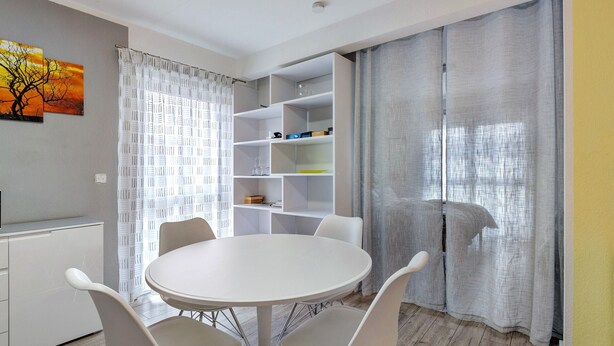 A modern room with a round white table, four chairs, a white shelving unit, and a large window with sheer curtains. - Apartment 'Haus Unterm Hohenstoffel' with Private Terrace and Wi-Fi (Hilzingen)