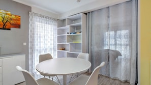 A modern room with a round white table, four chairs, a white shelving unit, and a large window with sheer curtains.