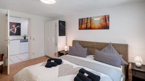 Ein Schlafzimmer mit einem Bett, Kissen, einem Nachttisch mit Lampe, einem Bild an der Wand und einer Tür zum Badezimmer.