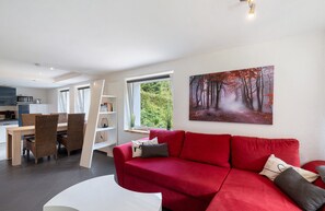A modern living room with a red sofa, a dining area with wicker chairs, and a kitchen in the background. - Apartment 'Langenwaldsee' with Private Terrace, Shared Garden and Wi-Fi (Freudenstadt)
