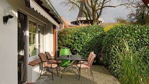 A patio with a table and chairs, a green trash bin, and a hedge.