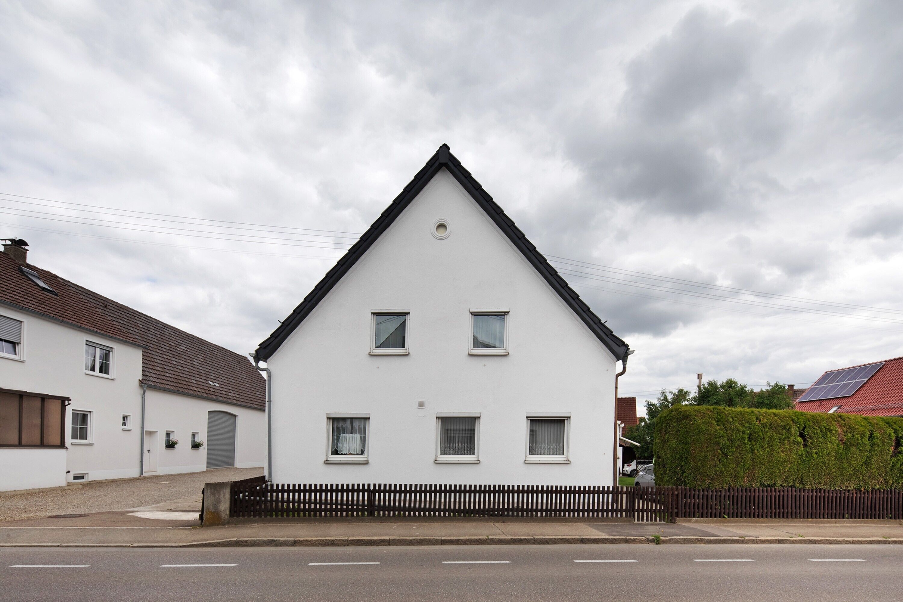 Een wit huis met twee verdiepingen, een puntig dak, een klein rond raam op de tweede verdieping en een zwarte omheining ervoor.