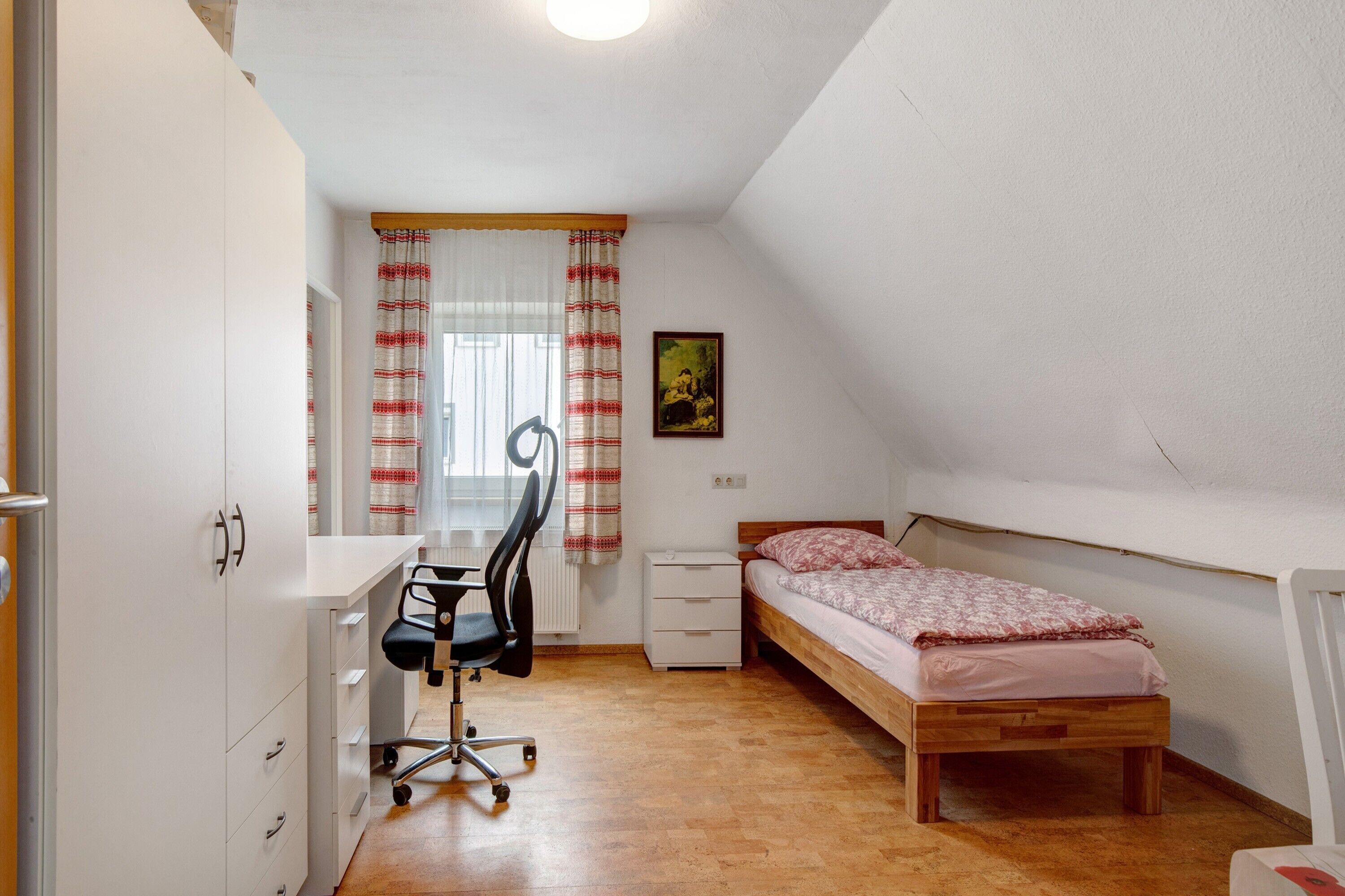 Een kleine, goed verlichte kamer met een houten bed, een bureau met stoel, een radiator en een raam met gordijnen.