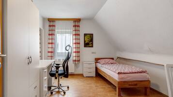 Een kleine, goed verlichte kamer met een houten bed, een bureau met stoel, een radiator en een raam met gordijnen.
