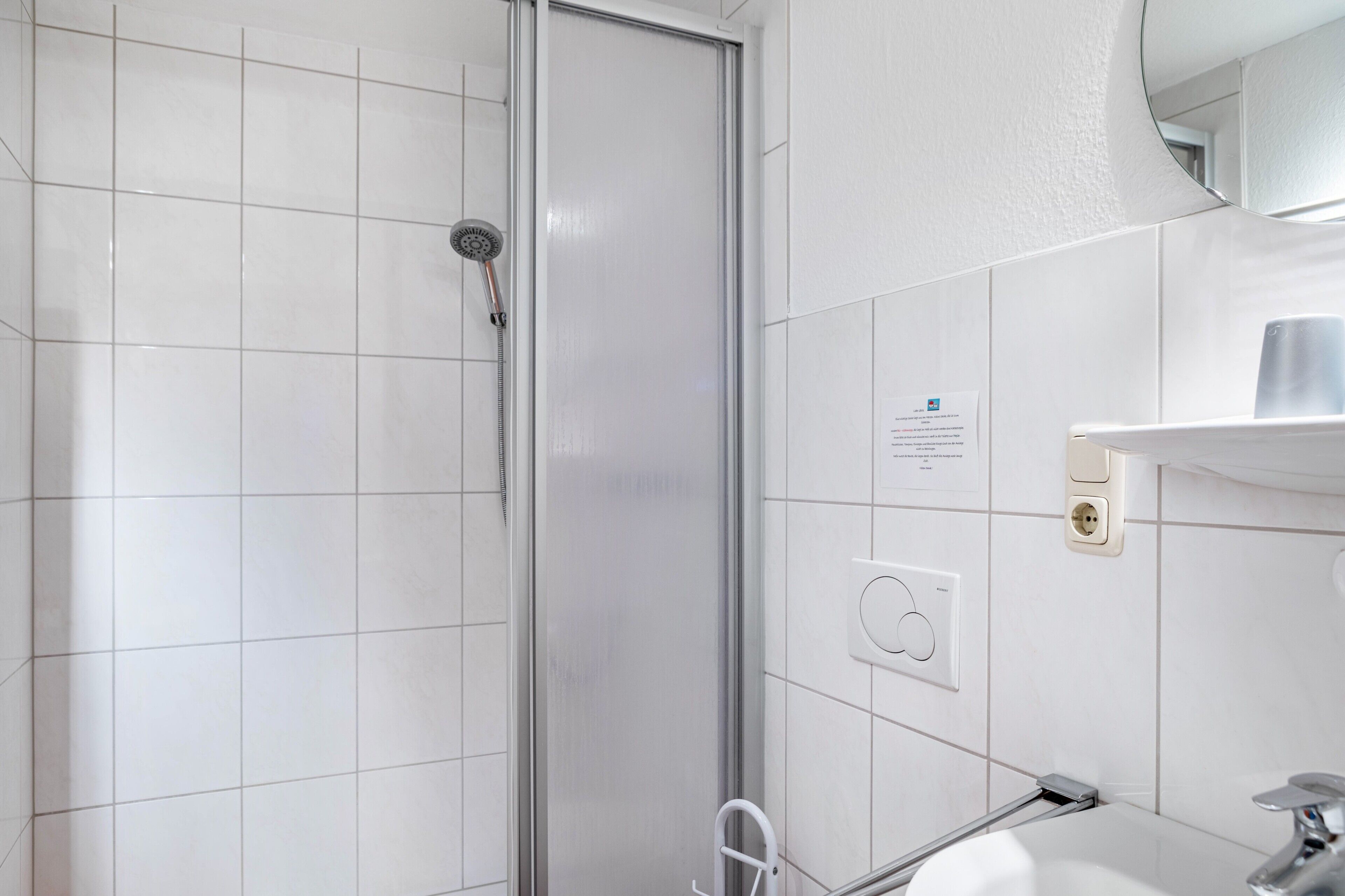 Moderni kylpyhuone, jossa on valkoiset laatat, lasinen suihkuseinä, pesuallas hanalla ja peili pesualtaan yläpuolella.