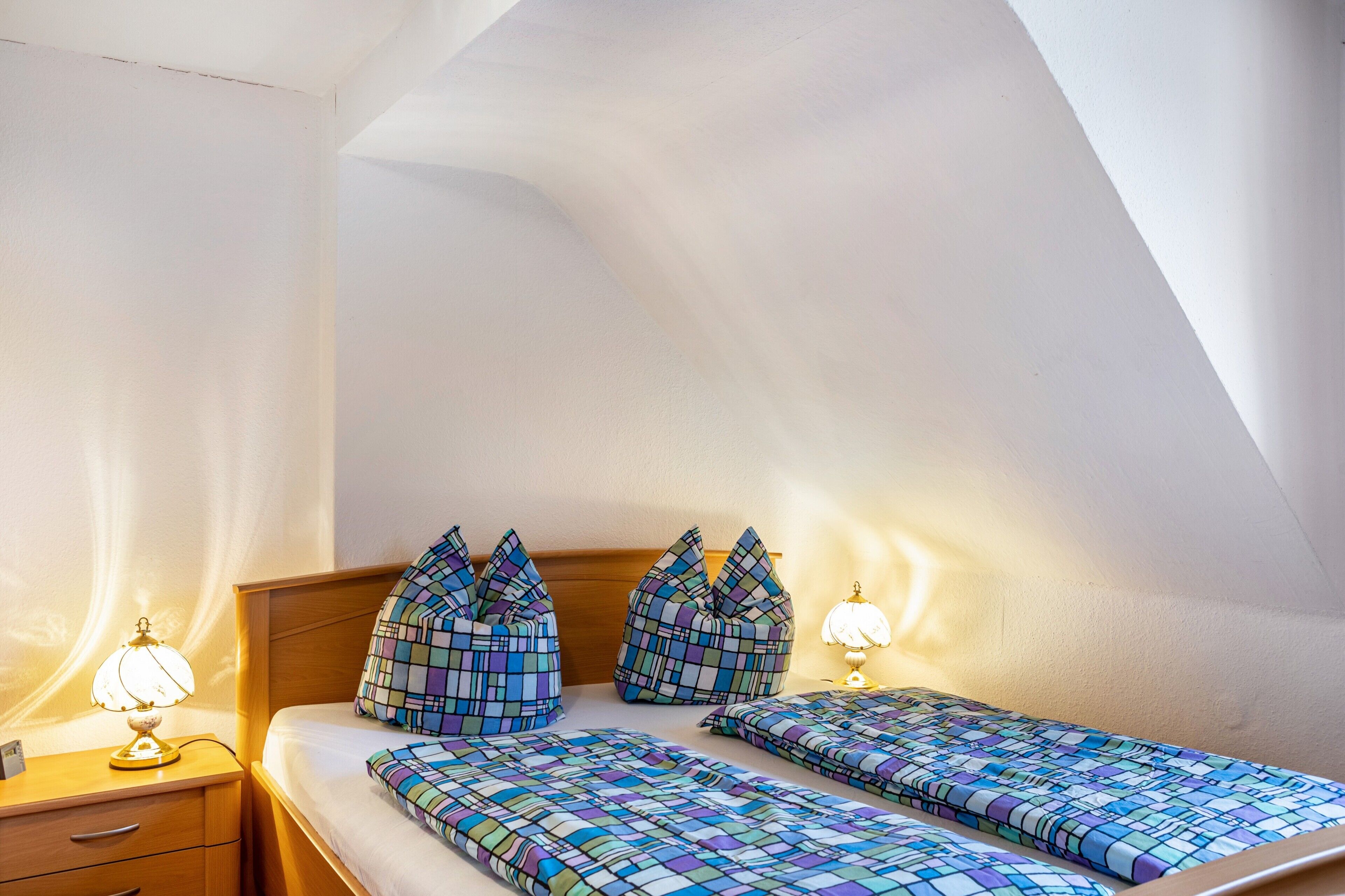 Um quarto com teto inclinado, duas camas com roupa de cama estampada, abajures e uma mesa de cabeceira com gavetas.