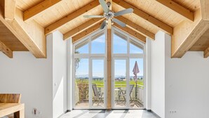 A room with a sloped wooden ceiling, a ceiling fan, large glass doors leading to an outdoor patio with chairs and a view of the countryside. - Holiday Apartment 'Cosy Home Allgäu' with Mountain View, Balcony and Wi-Fi (Haldenwang)
