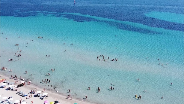 Una playa con agua turquesa cristalina, gente nadando y tomando el sol, y sombrillas de playa.