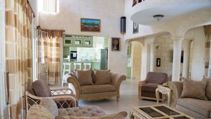 A spacious living room with a large sofa, armchairs, and a glass coffee table. A kitchen is visible in the background. - Marzalea - by Zenbreak (Paynes Bay)