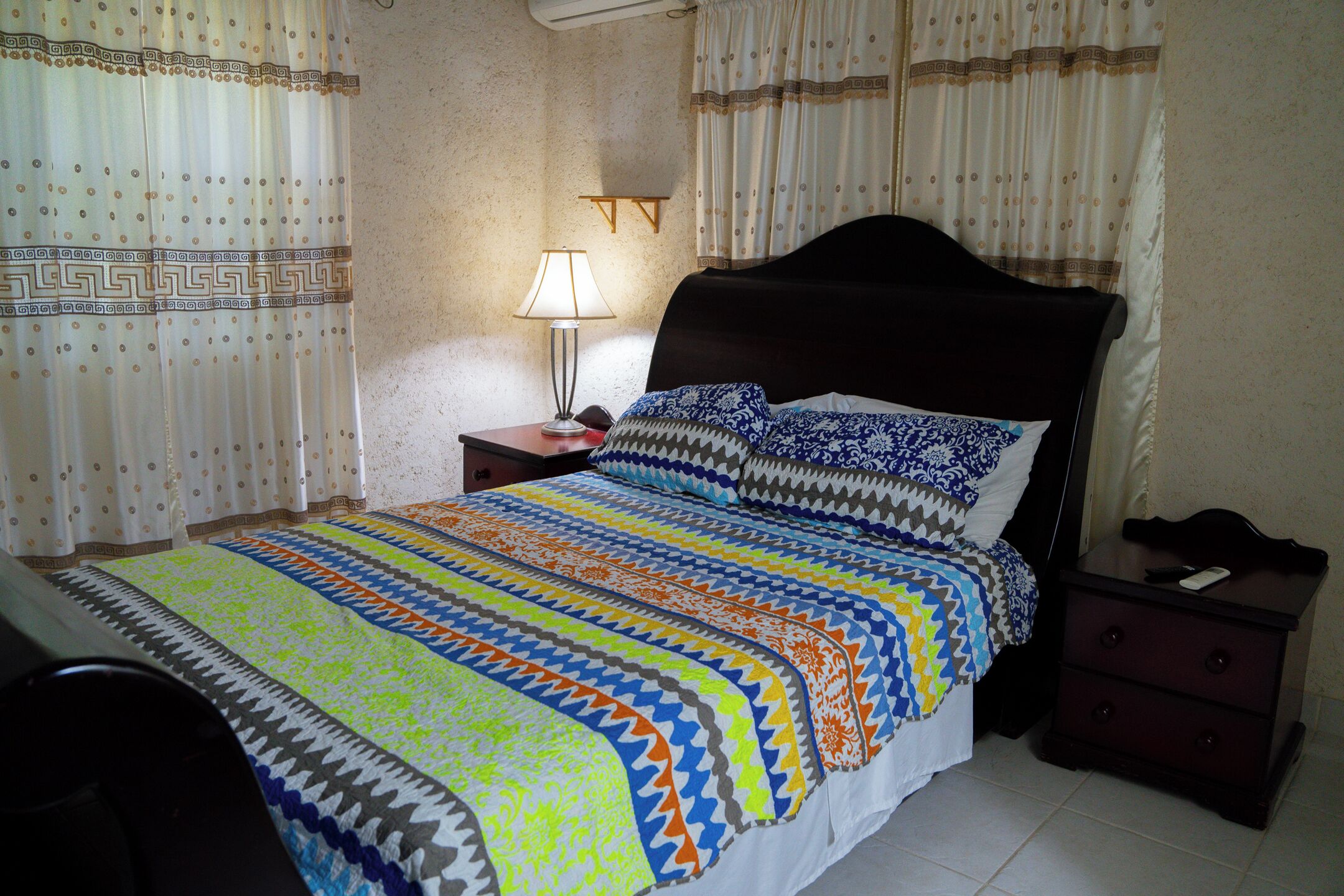 Una cama bien hecha con una colcha estampada, una mesita de noche con una lámpara y un pequeño mueble con cajones.