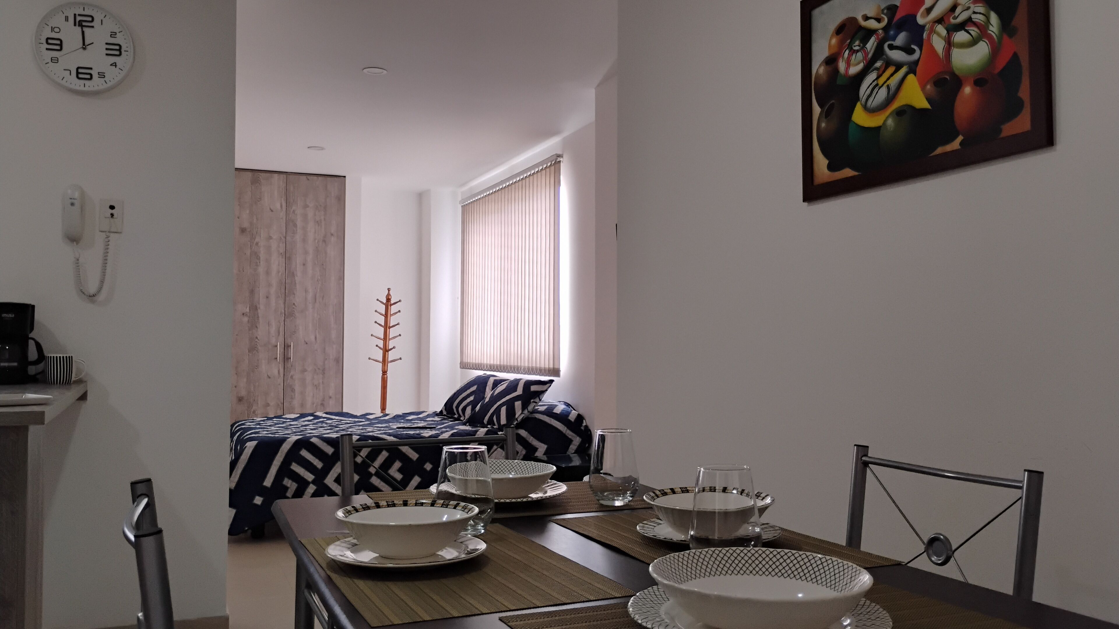 A modern hotel room with a dining area, a bed with patterned bedding, a painting on the wall, and a clock.