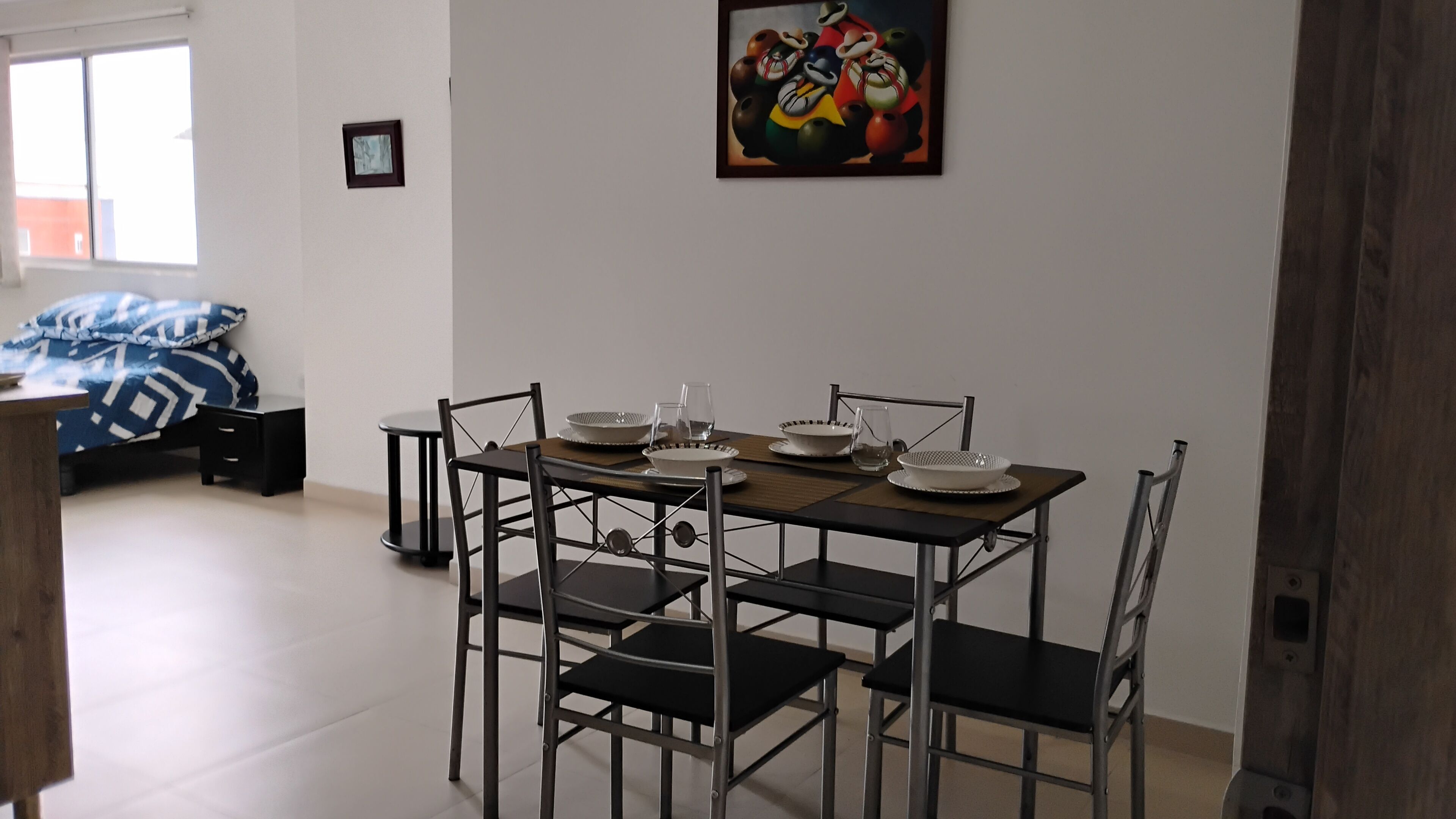 A dining area with a table set for four, surrounded by chairs, in a room with a bed and a painting on the wall.