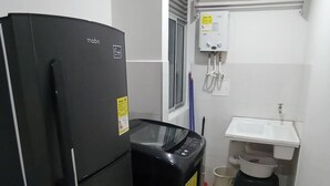 Una cocina compacta con un refrigerador negro, una lavadora negra, un lavamanos blanco y una ventana con persianas.