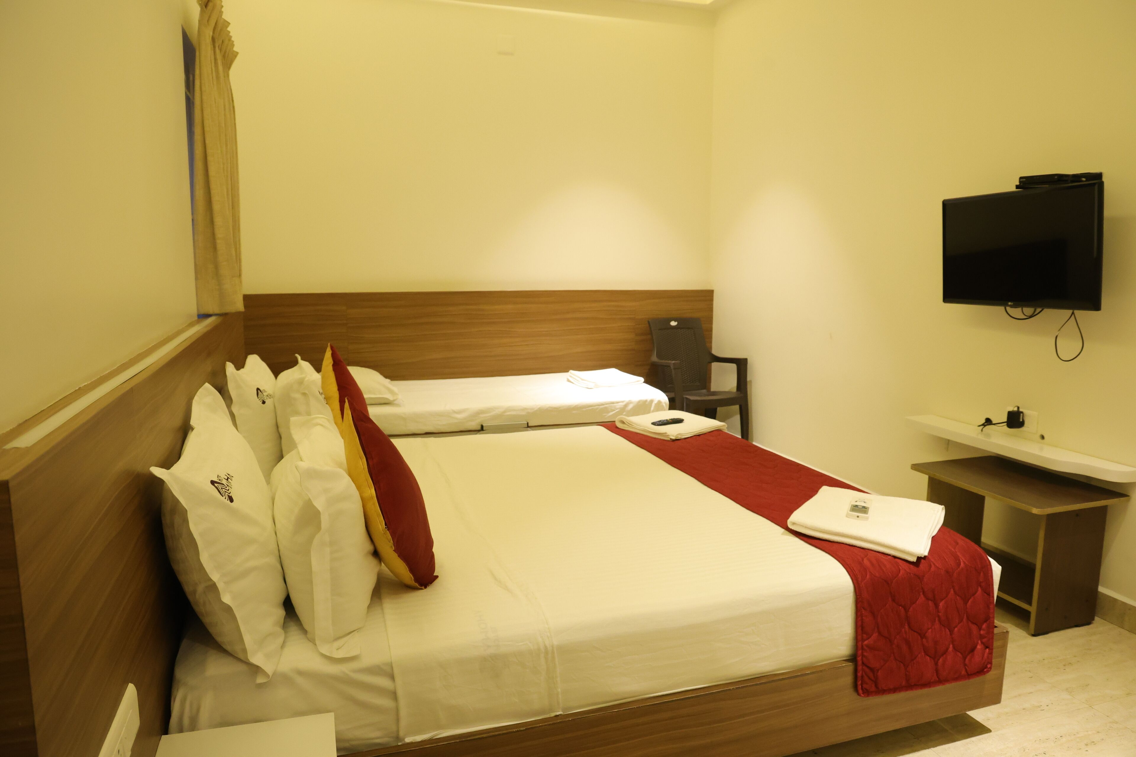A hotel room with a wooden headboard, a flat-screen TV mounted on the wall, a bedside table with a lamp, and a small desk with a sink.