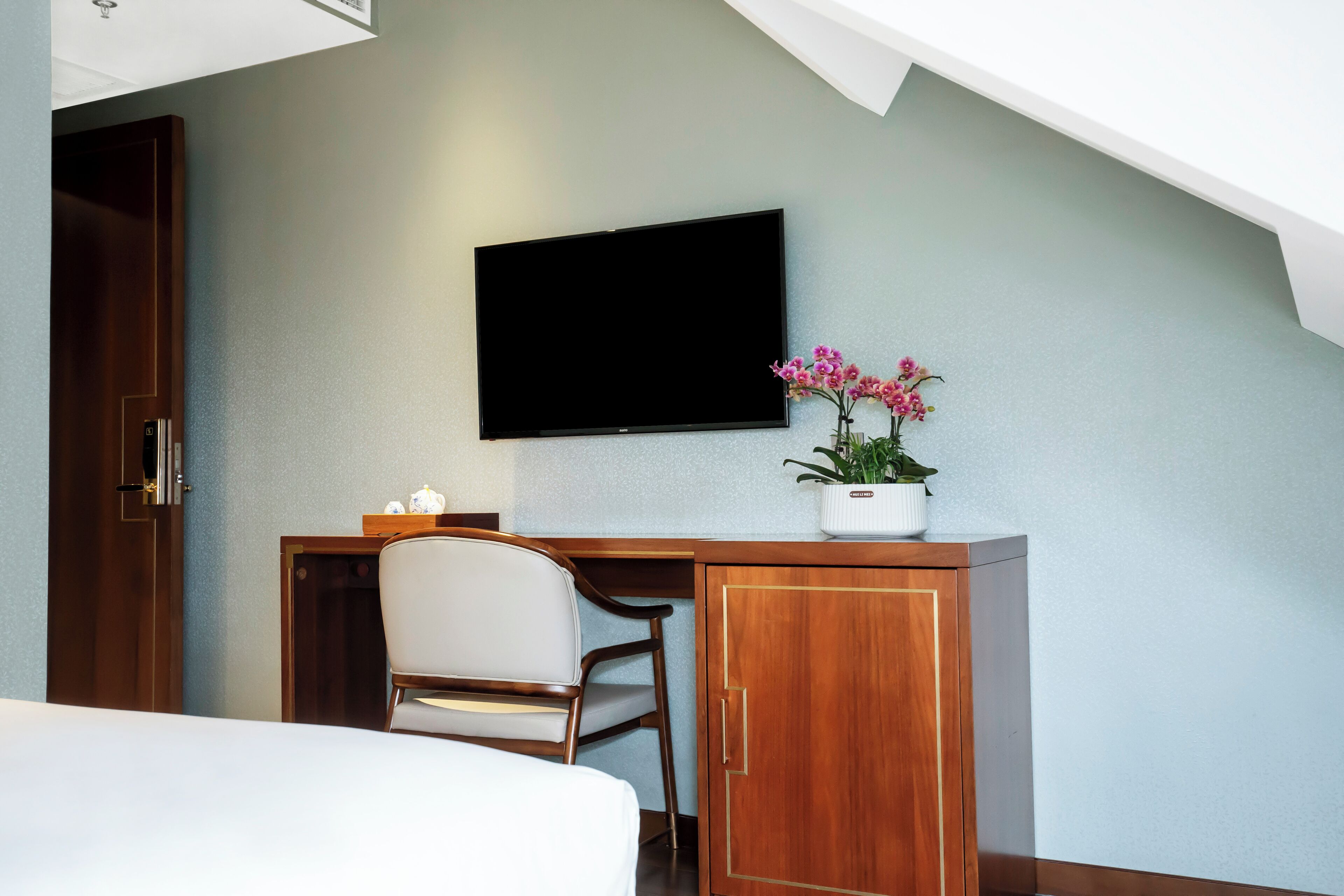 Une chambre d’hôtel comprenant un lit, un bureau avec une chaise, un téléviseur fixé au mur et une plante en pot.