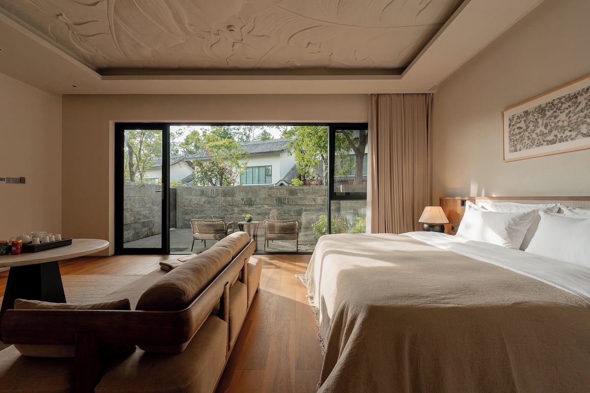 A modern hotel room with a large bed, a sofa, a dining table, and a view of the outside.