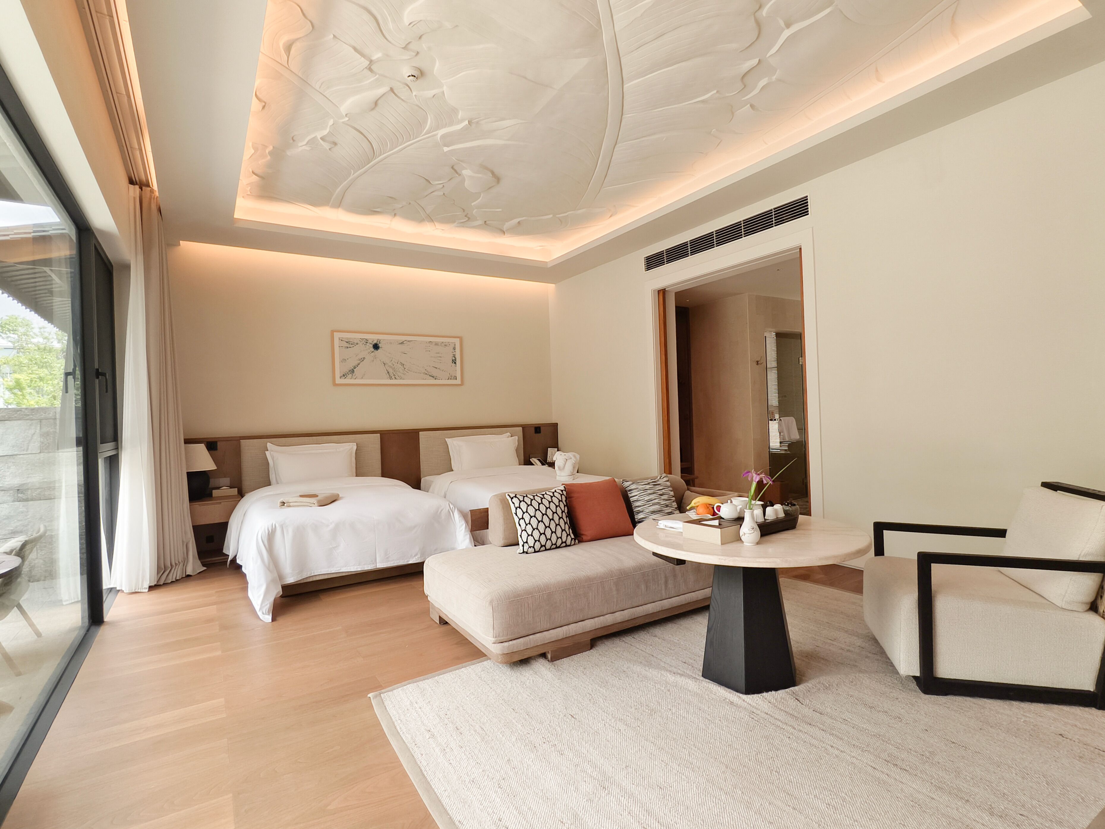 A hotel room with two beds, a round table, a sofa, a large mirror, and a view through a glass door.