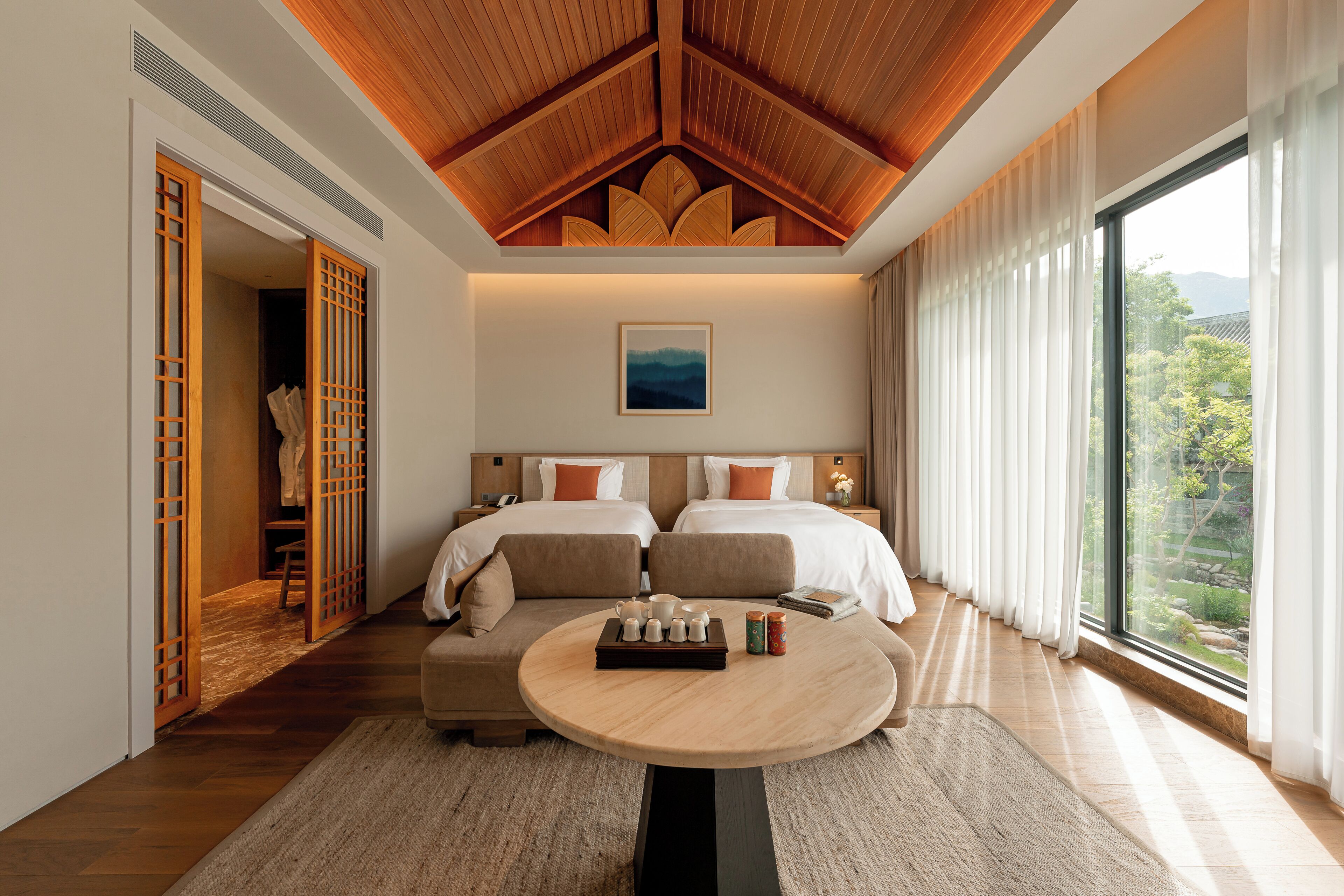 A hotel room with two beds, a round table with tea set, wooden ceiling, large window with view, and traditional sliding doors.